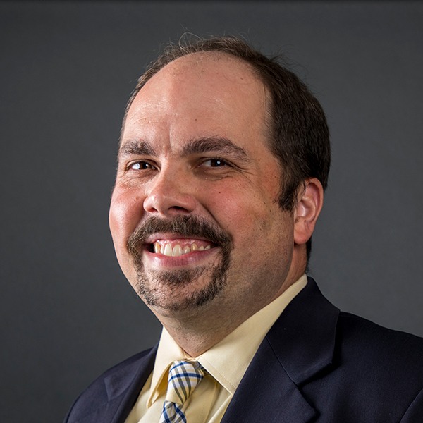 A man with short brown hair, a mustache, and a goatee, wearing a dark suit, light yellow shirt, and striped tie, smiles at the camera against a plain dark background.