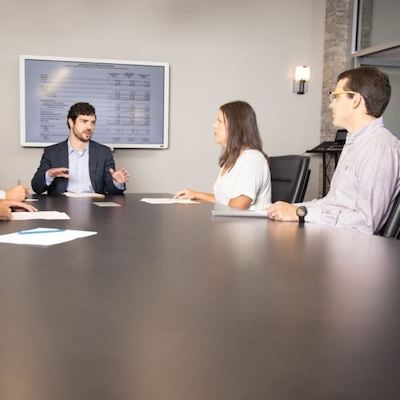 Four people sit around a conference table in a meeting room. One man is speaking while others listen. Papers are on the table, and a screen with charts and numbers is displayed on the wall behind them.