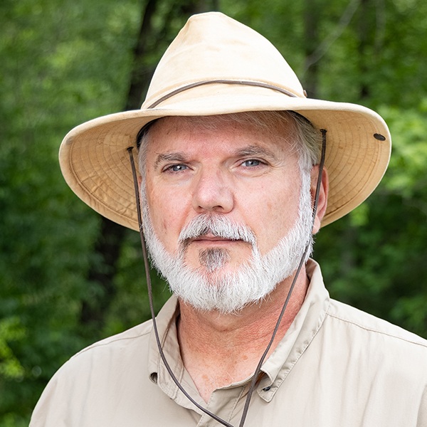 An older man with a white beard and mustache wears a beige safari hat and tan shirt, standing outdoors with green trees in the background.