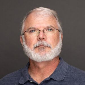 A middle-aged man with glasses, white hair, and a full white beard, wearing a blue collared shirt, poses in front of a plain gray background.