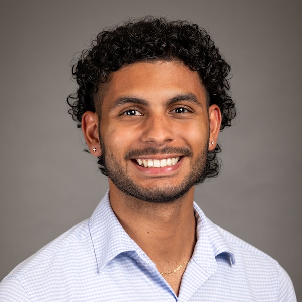A young man with curly dark hair and a beard, wearing a light blue button-down shirt, smiles at the camera in front of a plain dark gray background.
