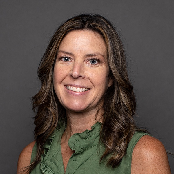 A woman with long wavy brown hair, wearing a sleeveless green blouse with ruffles, smiles in front of a plain dark gray background.