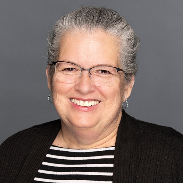 A smiling older woman with gray hair pulled back, wearing glasses, a black cardigan, and a black-and-white striped shirt, posed against a plain gray background.