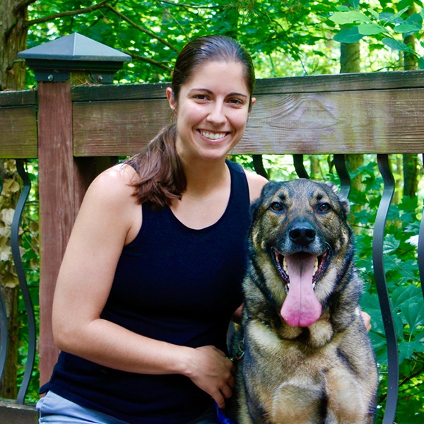 A woman in a black tank top smiles while kneeling next to a large, happy dog with its tongue out. They are outdoors, with green trees and a wooden railing in the background.