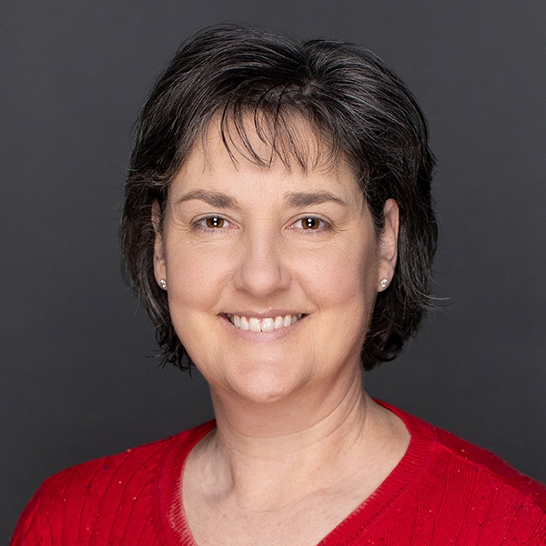 A woman with short dark hair and light skin smiles at the camera. She is wearing a red sweater and small stud earrings, with a plain dark gray background behind her.