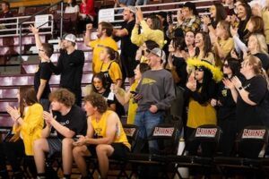 A lively group of fans wearing yellow and black cheer enthusiastically in bleachers at a sports event, with some standing and clapping while others sit and watch. Signs reading LIONS are visible on the seats.