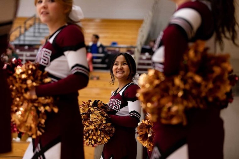 A cheerleader in a maroon and white uniform smiles while holding metallic pom-poms, surrounded by teammates. Bleachers and spectators are visible in the background.
