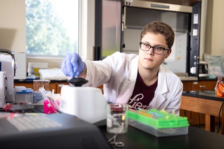 A young scientist wearing glasses and a lab coat works in a laboratory, handling equipment with a gloved hand. Various lab tools and colorful test tubes are arranged on the table.