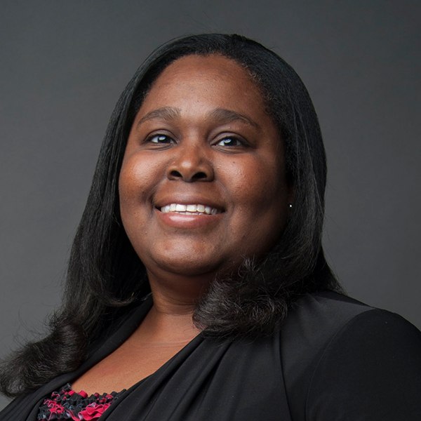 A woman with long dark hair smiles at the camera, wearing a black top and a black jacket, posing against a plain dark grey background.