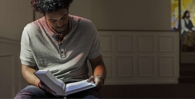 A man sits indoors reading a book, with sunlight illuminating the pages. In the background, a stained glass window featuring a religious figure is visible.