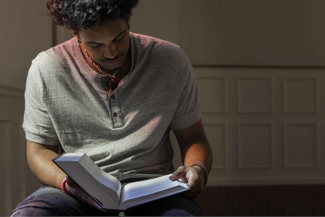 A young man with curly hair sits indoors, looking down and reading an open book in his lap. He wears a gray short-sleeve shirt and a bracelet, and is illuminated by soft, natural light.