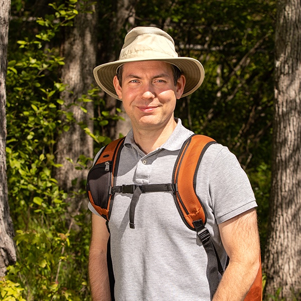 A man wearing a light gray polo shirt, a wide-brimmed hat, and an orange backpack stands outdoors in a sunlit forest, smiling at the camera.