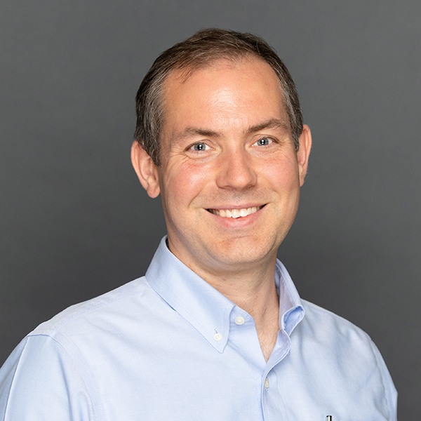 A man with short brown hair smiles at the camera, wearing a light blue button-up shirt. The background is plain and gray.