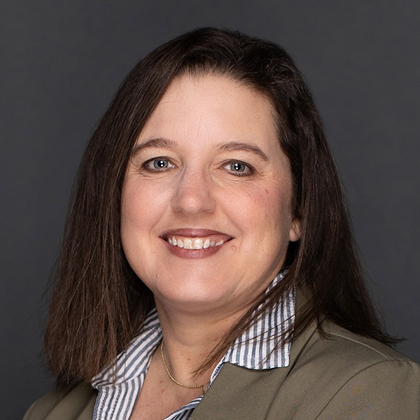 A woman with straight brown hair smiles at the camera. She is wearing a beige blazer over a blue and white striped shirt, with a simple gold necklace, against a plain dark background.