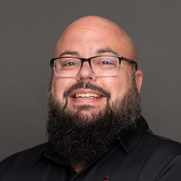 A smiling man with a bald head, full dark beard, and glasses wearing a black collared shirt, posed against a plain gray background.