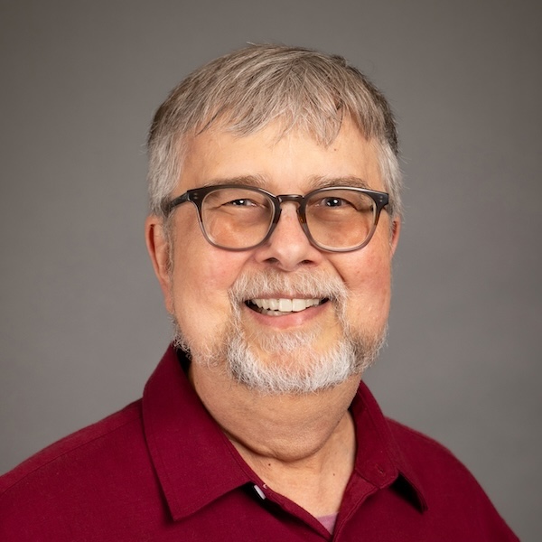 Smiling older man with gray hair, glasses, and a short beard, wearing a bright red shirt, posing against a plain dark gray background.