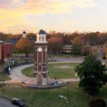 A clock tower stands in the center of a circular plaza surrounded by paths, grass, and trees, with campus buildings and a colorful sunset sky in the background.