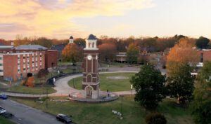 A clock tower stands in the center of a circular plaza surrounded by paths, grass, and trees, with campus buildings and a colorful sunset sky in the background.