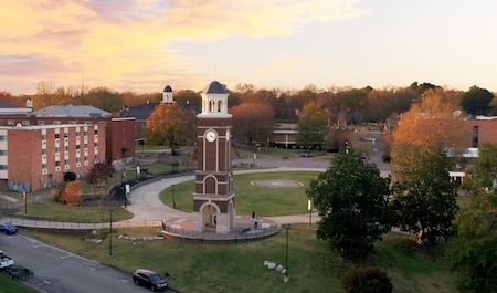 A tall brick clock tower stands in the center of a grassy campus quad, surrounded by trees and campus buildings under a colorful sunset sky. A curved path circles the tower, and a car is parked on a nearby street.