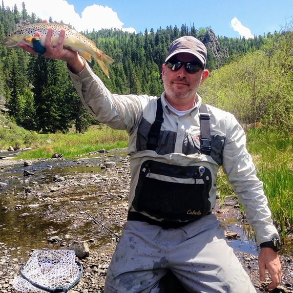 A man wearing sunglasses and fishing gear holds up a brown trout by a stream in a forested area on a sunny day. A net lies on the ground nearby.
