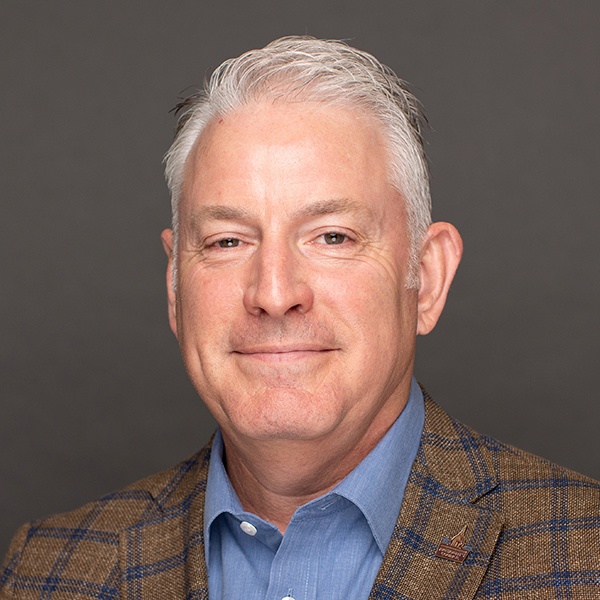A middle-aged man with short gray hair smiles softly. He is wearing a blue shirt and a brown plaid blazer, set against a plain gray background.