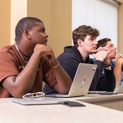 Three students sit at desks with laptops open, attentively listening and resting their chins on their hands in a classroom setting.
