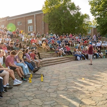 A large group of people sit on outdoor stone steps, listening to a person speaking in front of them on a sunny day. Trees and a brick building are visible in the background.