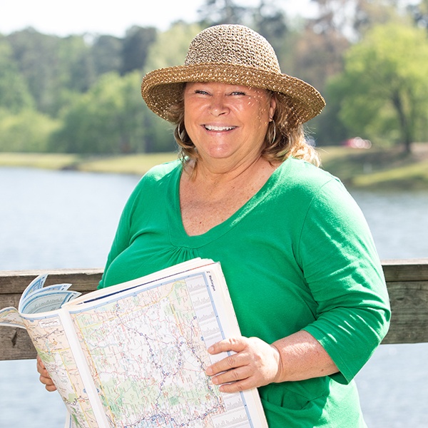 A woman in a green top and straw hat smiles while holding an open map. She is standing outside by a wooden railing with a lake and trees in the background on a sunny day.