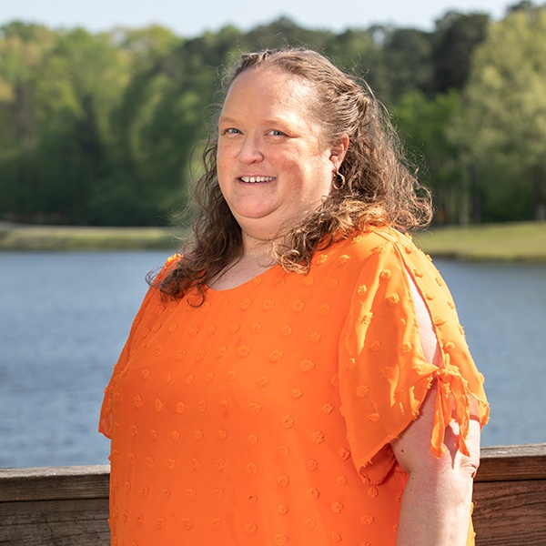 A woman with wavy brown hair wearing an orange short-sleeve top stands outdoors in front of a lake and trees, smiling at the camera.
