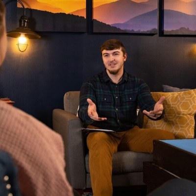 A young man sits on a couch in a cozy, warmly lit room, gesturing with his hands as he speaks to someone off camera. There are mountain-themed pictures and a yellow pillow in the background.