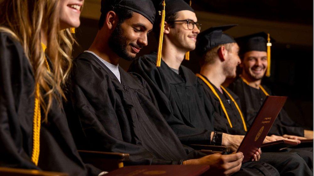 A group of graduates in black caps and gowns sit in a row, smiling and holding diplomas, as they celebrate their graduation ceremony.