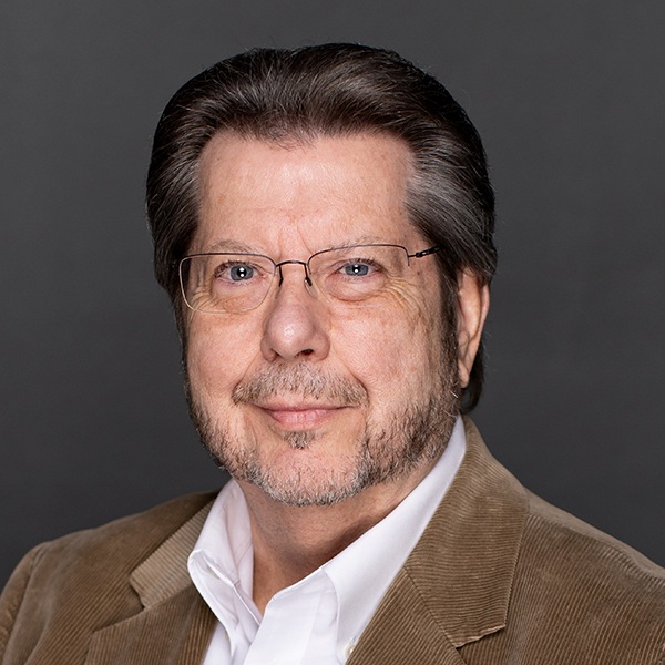A middle-aged man with glasses, short brown hair, and a trimmed beard, wearing a tan corduroy jacket over a white shirt, poses in front of a plain dark gray background.