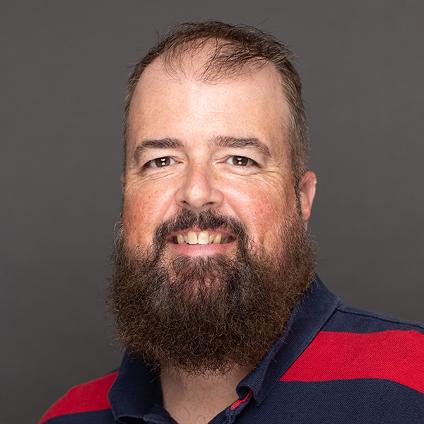 A bearded man with short hair smiles at the camera. He is wearing a navy blue and red striped polo shirt, and the background is plain and gray.