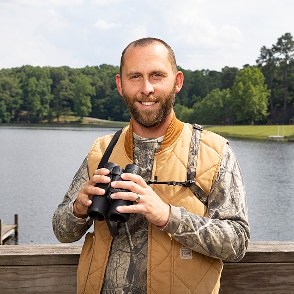 A smiling man in a camouflage shirt and tan vest holds binoculars while standing by a lake with trees in the background on a partly cloudy day.