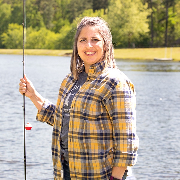 A woman wearing a yellow and blue plaid shirt stands by a lake, smiling and holding a fishing pole. Trees and greenery are visible in the background on a sunny day.