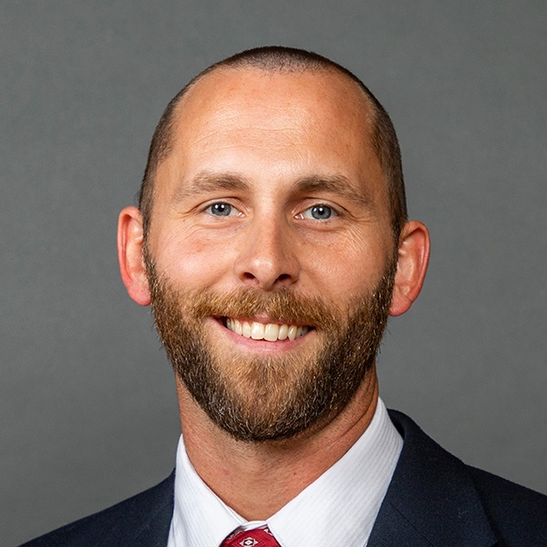 A smiling man with a trimmed beard and short hair, wearing a dark suit, white shirt, and red patterned tie, posed in front of a plain gray background.