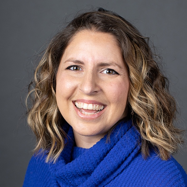 A woman with wavy, shoulder-length light brown hair is smiling. She is wearing a bright blue sweater and is posed in front of a plain gray background.
