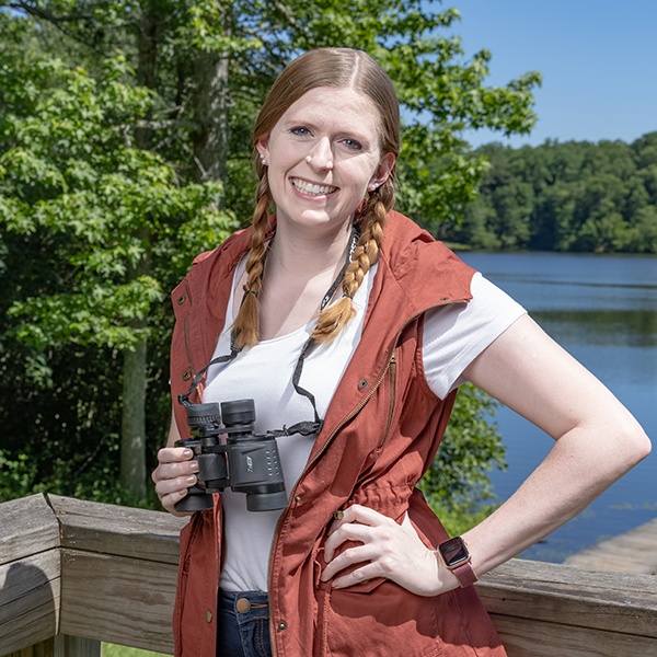 A smiling woman with braided hair stands on a wooden deck by a lake, holding binoculars. She wears a rust-colored vest over a white shirt, and there are green trees and blue sky in the background.