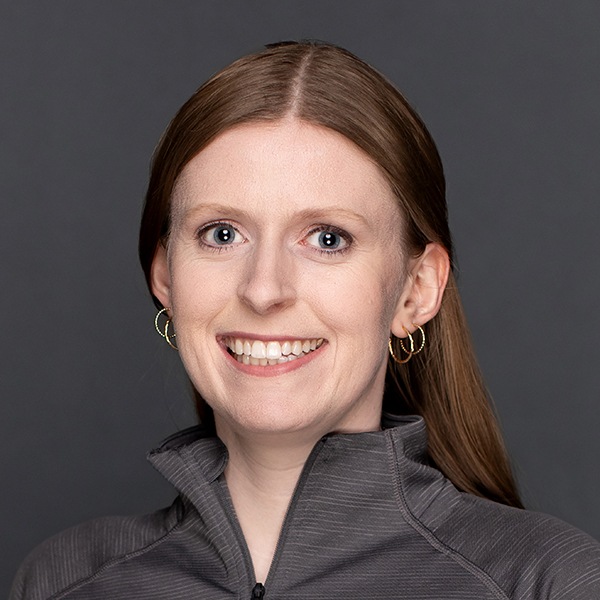 A woman with straight, light brown hair and hoop earrings smiles at the camera. She is wearing a gray zip-up top and is posed against a plain dark gray background.