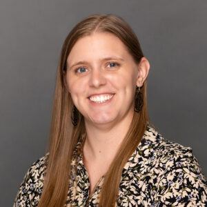 A woman with long straight light brown hair, wearing a patterned blouse and black earrings, smiles at the camera against a plain gray background.