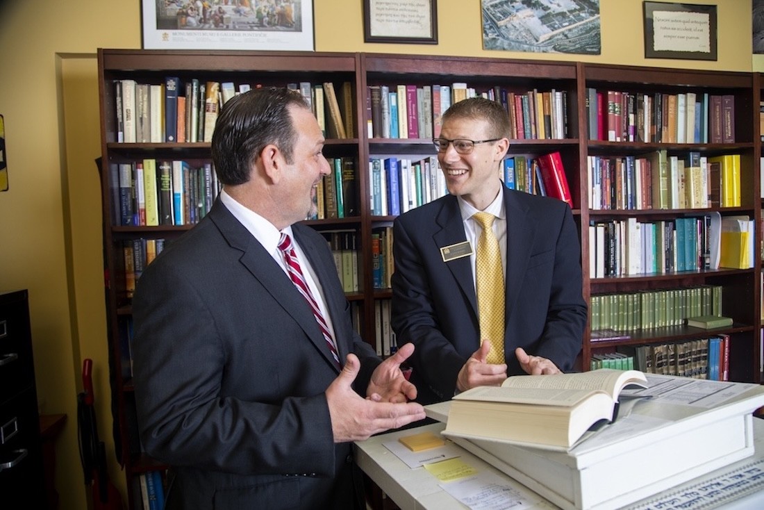 Two men in suits are having a conversation and smiling in an office with shelves of books behind them. Open books and papers are spread out on the desk in front of them.