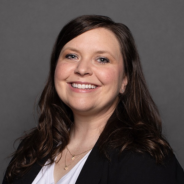 A woman with long brown hair smiles at the camera. She is wearing a black blazer over a white top and has layered necklaces. The background is plain and gray.