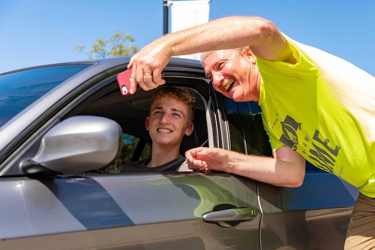 An older man in a yellow shirt smiles and takes a selfie with a younger man who is sitting in the drivers seat of a gray car on a sunny day.