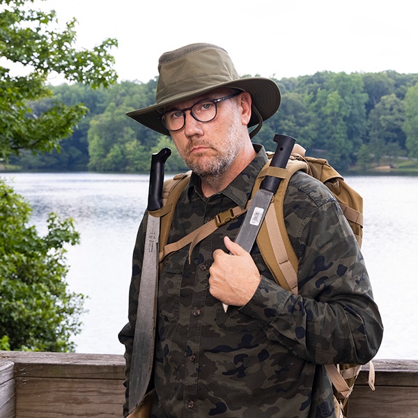 A man in glasses and a wide-brimmed hat, wearing a camouflage shirt and backpack, stands outdoors near a lake, holding a large machete with another machete strapped to his bag. Trees and water are visible in the background.