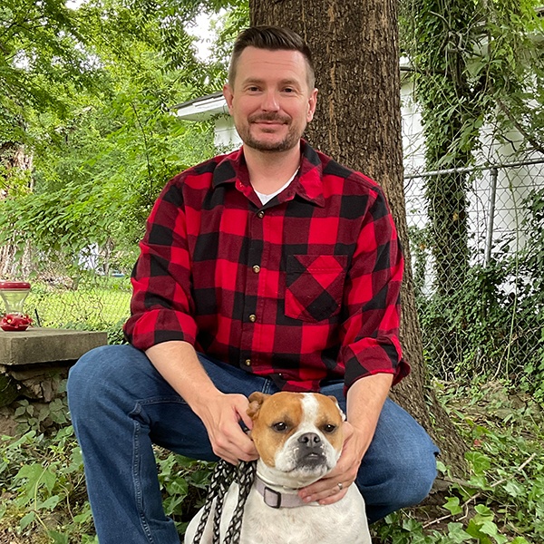 A man in a red plaid shirt sits outside on a stone ledge, smiling at the camera while petting a brown and white dog. Green trees and a chain-link fence are visible in the background.