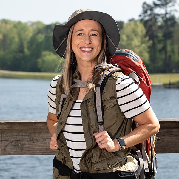 A woman wearing a wide-brim hat, striped shirt, and vest stands outdoors on a wooden bridge with a red hiking backpack, smiling with a lake and trees in the background.
