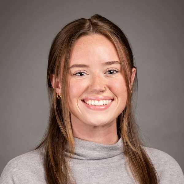 A young woman with long brown hair smiles at the camera. She is wearing a black top, gold hoop earrings, a gold necklace, and has a small nose ring. The background is plain and dark gray.