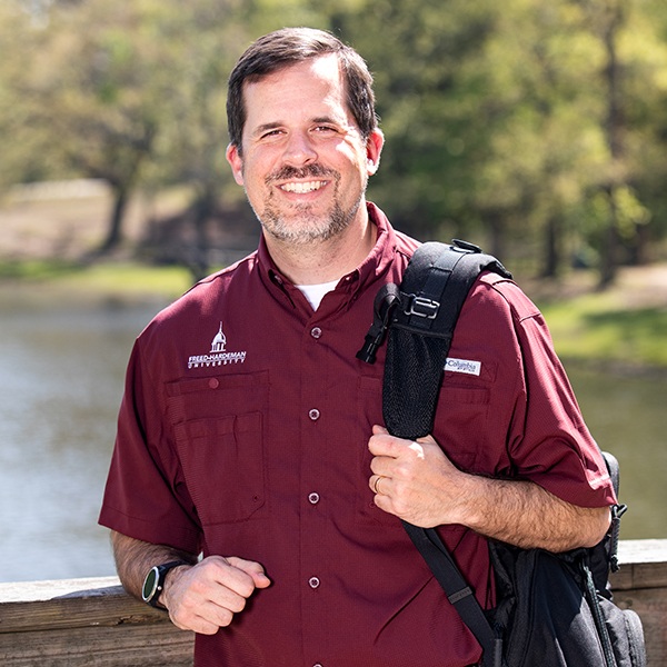 A man with short dark hair and a beard, wearing a maroon button-up shirt and a backpack, stands smiling on a wooden bridge with water and trees in the background on a sunny day.