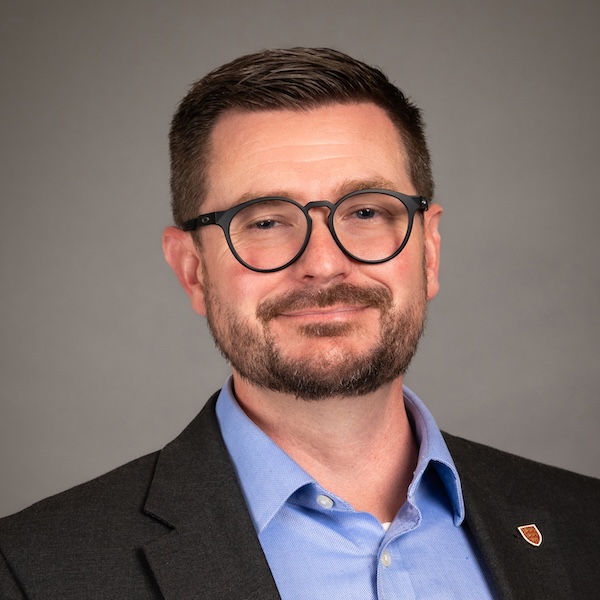 A man with short dark hair, glasses, and a trimmed beard is wearing a dark checkered blazer, a navy blue shirt with small white dots, and a red patterned tie, posing against a dark gray background.