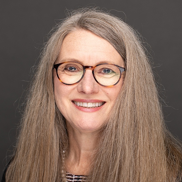 Smiling woman with long gray hair, wearing round eyeglasses and a black top, posed in front of a plain dark background.